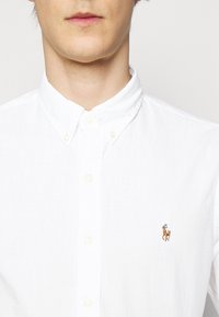 Bílá košile s klasickým límcem, s malým vyšitým hnědým logem koně na levé straně hrudi a texturovanou látkou.