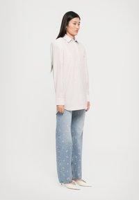Mulher vestindo uma camisa de mangas largas listrada em rosa claro, jeans azuis com estampas florais em rosa e sapatos de salto kitten brancos com bico fino, contra um fundo branco.