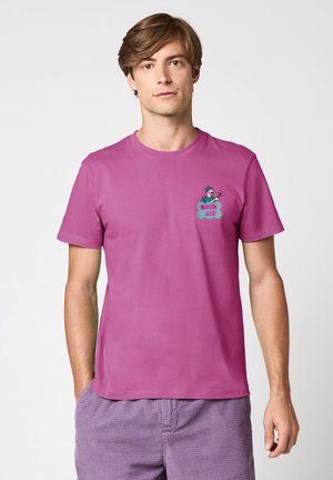 Jeune homme portant un T-shirt violet avec un motif brodé coloré "Open Air" et un short violet texturé, debout devant un fond uni.