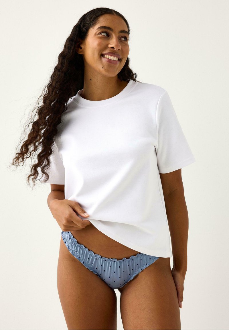 T-shirt blanc à manches courtes ; coupe décontractée, tissu lisse. Culotte bleue claire à volants avec pois marine. Modèle tenant le t-shirt d'une main.