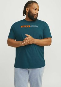 Teal kortærmet T-shirt med "JACK & JONES" logo i orange og hvid. Blødt bomuldsstof, afslappet pasform, parret med lyseblå jeans.