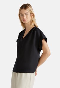 Blouse noire à manches courtes, avec un col en V, en tissu texturé et à coupe décontractée. Design simple sans motifs visibles ni accessoires.