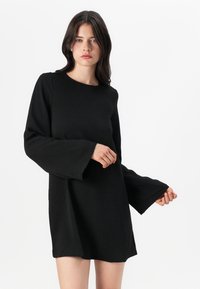 Vestido negro con mangas largas y acampanadas, escote redondo y un corte holgado. La tela parece texturizada, ofreciendo un aspecto suave y confortable.