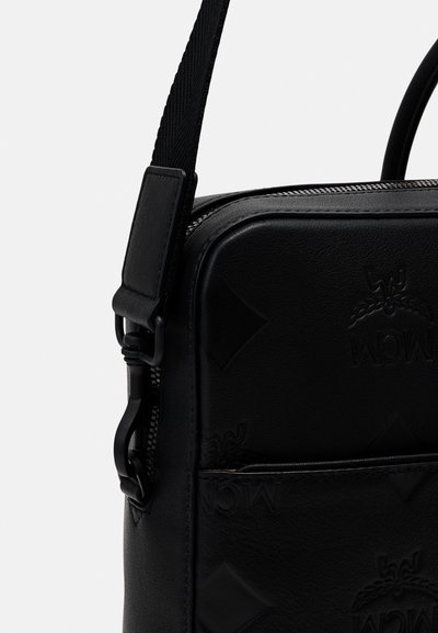 Sac en cuir noir avec une finition texturée, logo embossé, fermeture zippée et une bandoulière amovible. Forme rectangulaire compacte avec des accents minimaux.