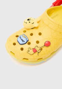 Sabot jaune avec des breloques à thème Winnie l'Ourson : une tête en peluche, un pot de miel et Winnie tenant un ballon rouge.
