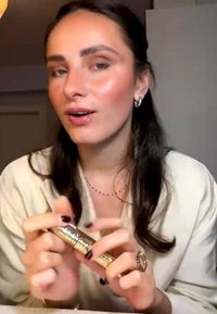 Giovane donna con capelli scuri tiene e dimostra un prodotto cosmetico dorato, indossando una maglietta bianca e gioielli minimalisti in casa.