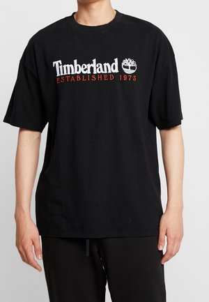 Osoba nosząca czarną koszulkę Timberland z białym logo i czerwonym napisem "Established 1973", w połączeniu z czarnymi spodniami na jednolitym tle.