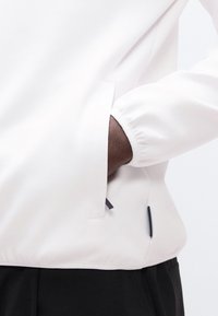 Άτομο που φοράει λευκό σακάκι με το χέρι στην πλαϊνή τσέπη, συνδυασμένο με μαύρο παντελόνι, δείχνοντας τις λεπτομέρειες της μανσέτας και της τσέπης του σακακιού.