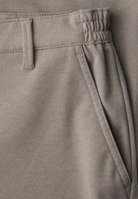 Détail d'un pantalon en tissu beige montrant la ceinture élastique, les passants de ceinture et une poche latérale inclinée avec des coutures visibles.