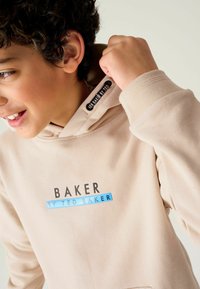 Sudadera con capucha beige que presenta un gráfico frontal con la palabra "BAKER" en negro y "by TED BAKER" en azul. Tejido suave y puños acanalados.