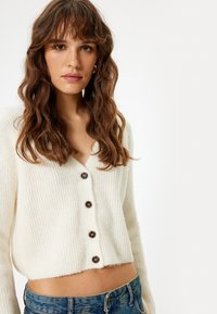 Weißer gerippter Cardigan mit V-Ausschnitt, ausgestattet mit dunklen Knöpfen und einem cropped Design, getragen über blauen Jeans. Weiche Textur.