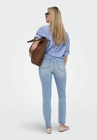 Femme blonde portant des lunettes de soleil, une chemise rayée bleue, un jean clair, des ballerines beiges, portant un grand sac en cuir marron sur l'épaule, de dos.
