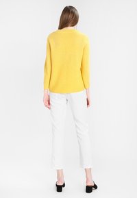 Gele, gestructureerde trui met driekwartsmouwen gecombineerd met witte broek met fijne strepen en zwarte schoenen met blokhak, gezien van achteren.