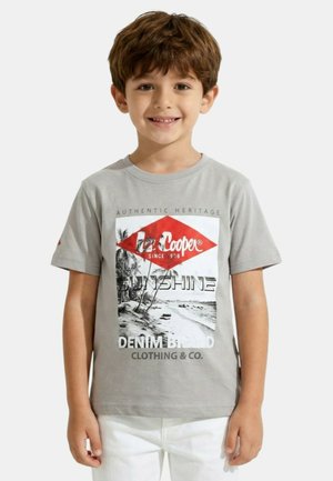 Jeune garçon souriant aux cheveux bruns portant un t-shirt graphique gris Lee Cooper et un pantalon blanc, debout devant un fond uni.