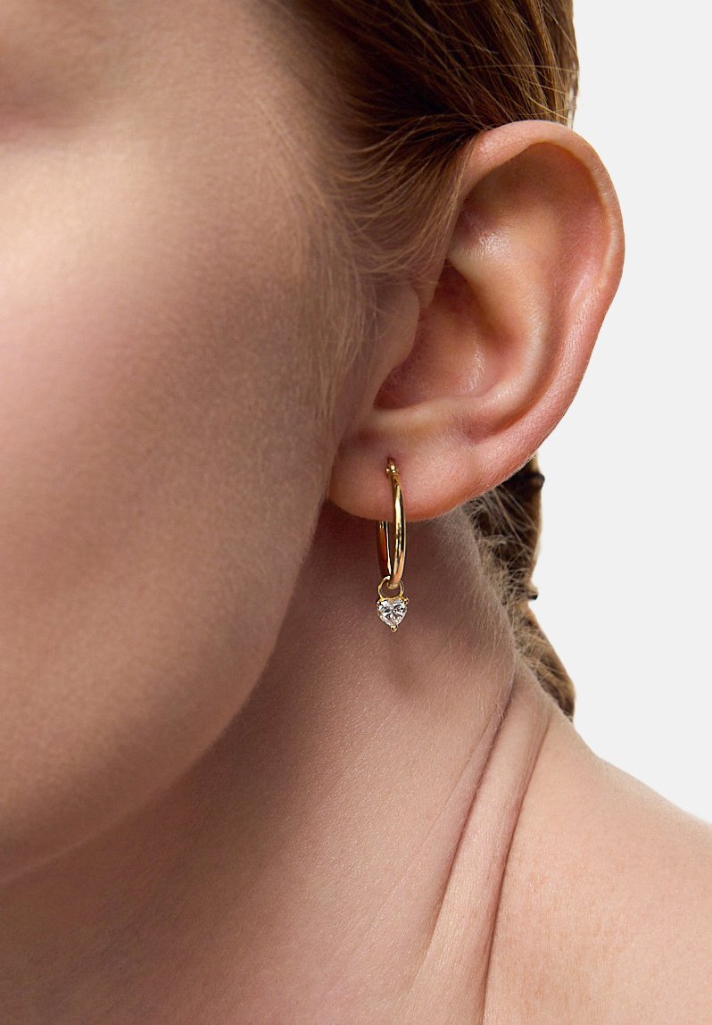 Złoty kolczyk w formie obręczy z zawieszką w kształcie serca, wykonaną z przezroczystego kamienia szlachetnego, o gładkiej teksturze i błyszczącym wykończeniu, zawieszony na małym zapięciu.