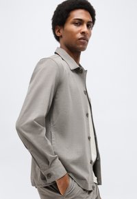 Mand med kort, krøllet hår iført en lysegrå jakke med knapper og matchende bukser, stående med den ene hånd i lommen mod en hvid baggrund.