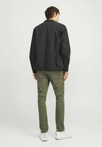 Camicia nera con bottoni e maniche lunghe, abbinata a pantaloni verde oliva e sneakers bianche. Il tessuto sembra liscio e leggero.
