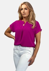 Fuksjowa bluzka z kołnierzykiem mandarynkowym, krótkimi rękawami i złotym guzikiem przy dekolcie. Połączona z białymi, dopasowanymi spodniami. Gładka tekstura.