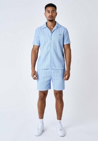 Camicetta a maniche corte a righe blu abbinata a pantaloni corti coordinati. Il tessuto è leggero, con chiusura a bottoni e una vestibilità rilassata. Sneakers bianche completano il look.