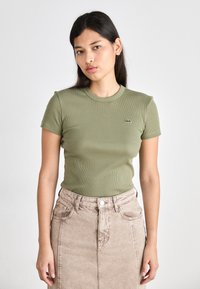 Olive green ribbed short-sleeve t-shirt with a fitted design, displayed with a light beige denim skirt featuring front buttons and pockets.