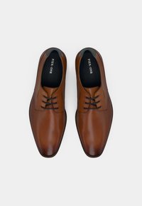 Un paio di scarpe eleganti in pelle marrone lucida con lacci neri e fodera interna nera, posizionate lato a lato su uno sfondo bianco.