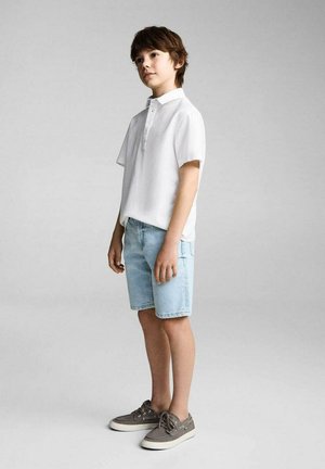 Polo shirt blanc à manches courtes, shorts en denim bleu clair, baskets en toile grises avec semelles blanches, se tenant sur un fond neutre.