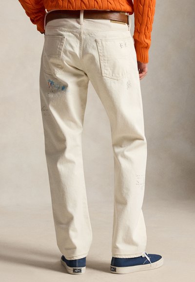 Person som har på seg off-white jeans med subtile trykte mønstre, et brunt belte, oransje genser og blå joggesko, stående mot en ensfarget bakgrunn.