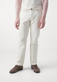 Jeanși din denim alb, cu croială slim, având un design clasic cu cinci buzunare și închidere cu buton de sus, asortați cu adidași din suede maro.