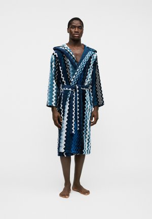 Homme portant un peignoir à capuche mi-long avec des motifs en zigzag bleus et blancs, debout pieds nus devant un fond uni.