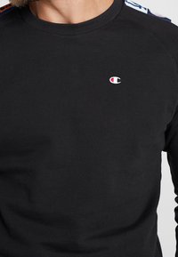 Svart sweatshirt i bomullsblandning med ribbad rund halsringning, som har en liten broderad logotyp på bröstet och mönstrade detaljer på axlarna.