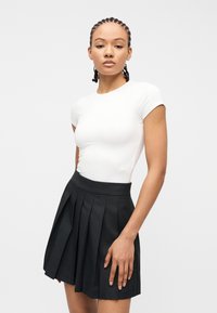 Νεαρή γυναίκα με πλεξούδες, ντυμένη με εφαρμοστό λευκό κοντομάνικο τοπ και μαύρη πλισέ φούστα, στέκεται μπροστά σε ένα απλό ανοιχτόχρωμο φόντο.