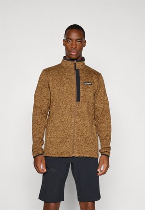 Veste en polaire marron avec col montant, fermeture éclair sur toute la longueur à l'avant et logo sur la poitrine, associée à un short foncé, sur fond uni.