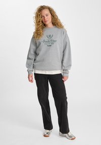Sudadera gris con logo y texto en verde oscuro, de corte holgado, con puños acanalados, llevada sobre una camisa blanca, combinada con pantalones negros y zapatos plateados.