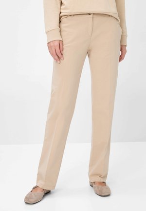 Person wearing beige straight-leg pants and beige long-sleeve top, standing with feet slightly apart, beige flats visible.