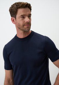 Granatowy sweter z krótkim rękawem wykonany z miękkiego materiału. Posiada ribbowany okrągły dekolt oraz dopasowany krój. Gładka, jednolita tekstura.