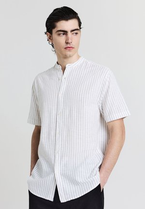 Giovane uomo che indossa una camicia bianca a maniche corte con sottili righe verticali nere, con le mani in tasca, che guarda di lato.