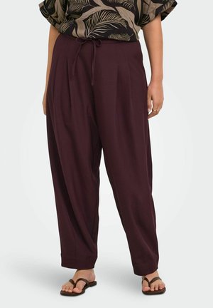 Femme portant un pantalon ample violet foncé avec une ceinture à cordon et des sandales marron sur un fond uni.