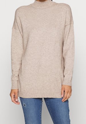 Personne portant un pull en maille beige à manches longues et col haut, associé à un jean bleu usé, debout devant un fond clair uni.