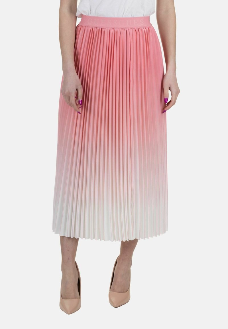Femme portant une jupe midi plissée dégradée du rose au blanc, un haut blanc et des talons hauts beiges, debout devant un fond uni.