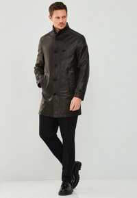 Bruine leren pea coat met een staande kraag, dubbelzijdige knopen, twee zijzakken en een gestructureerde afwerking. Gemaakt over zwarte broek en schoenen.