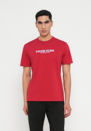Punane puuvillane T-särk lühikeste varrukatega, millel on valge logo tekst "CALVIN KLEIN ESTABLISHED 1968" rinna kohal. Sile tekstuur.