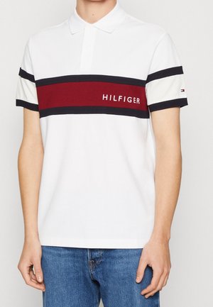 Valkoinen polo paita, jossa on punaisia ja laivastonsinisiä vaakasuoria raitoja, ja jossa lukee "HILFIGER" brodeerattuna rinnassa. Puuvillamateriaali, lyhyet hihat.