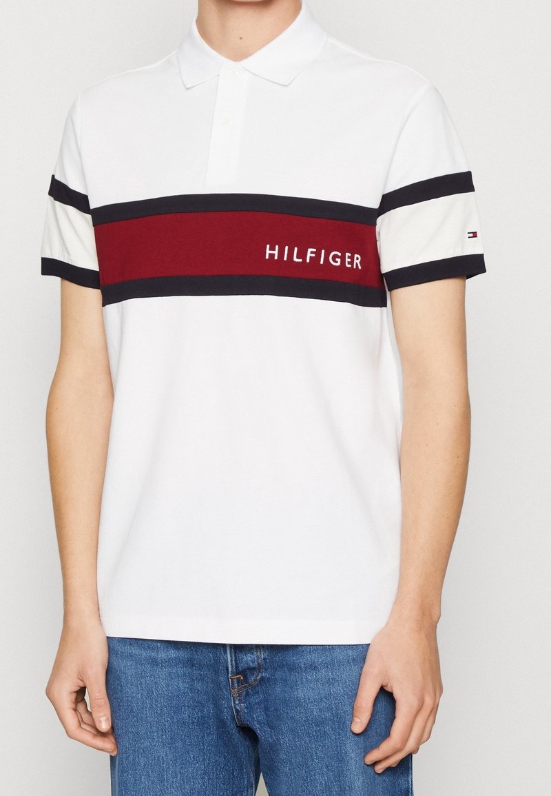 Valkoinen polo paita, jossa on punaisia ja laivastonsinisiä vaakasuoria raitoja, ja jossa lukee "HILFIGER" brodeerattuna rinnassa. Puuvillamateriaali, lyhyet hihat.