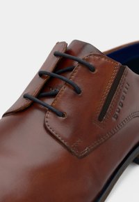 Rjava usnjena čevlja s črnimi vezalkami, s gladko teksturo, všitimi poudarki in subtilnim logotipom blagovne znamke na strani.