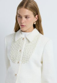 Chaqueta de tweed blanca con embellishments de perlas en el frente, que presenta un cuello clásico y botones con acentos dorados.