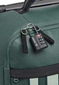 Borsa in tessuto verde con superficie testurizzata, dotata di lucchetto combinato, zip e dettagli in webbing neri. Brandizzazione sottile visibile.