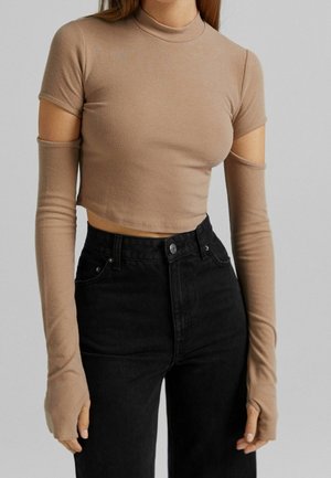 Femme portant un top court beige avec manches longues détachables et un jean noir taille haute sur un fond gris uni.