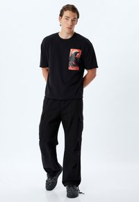 Camiseta de algodón negra con un diseño gráfico en el bolsillo frontal que presenta tonos naranjas y negros, combinado con pantalones cargo negros y zapatillas oscuras.