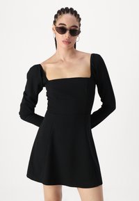 Vestido negro de manga larga con escote cuadrado, cuerpo texturizado y falda acampanada. Combinado con gafas de sol negras y pendientes de aro.