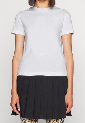 Kvinde iført en enkel hvid T-shirt med et subtilt logo på brystet, kombineret med et sort plisseret skørt og mønstrede leggings.